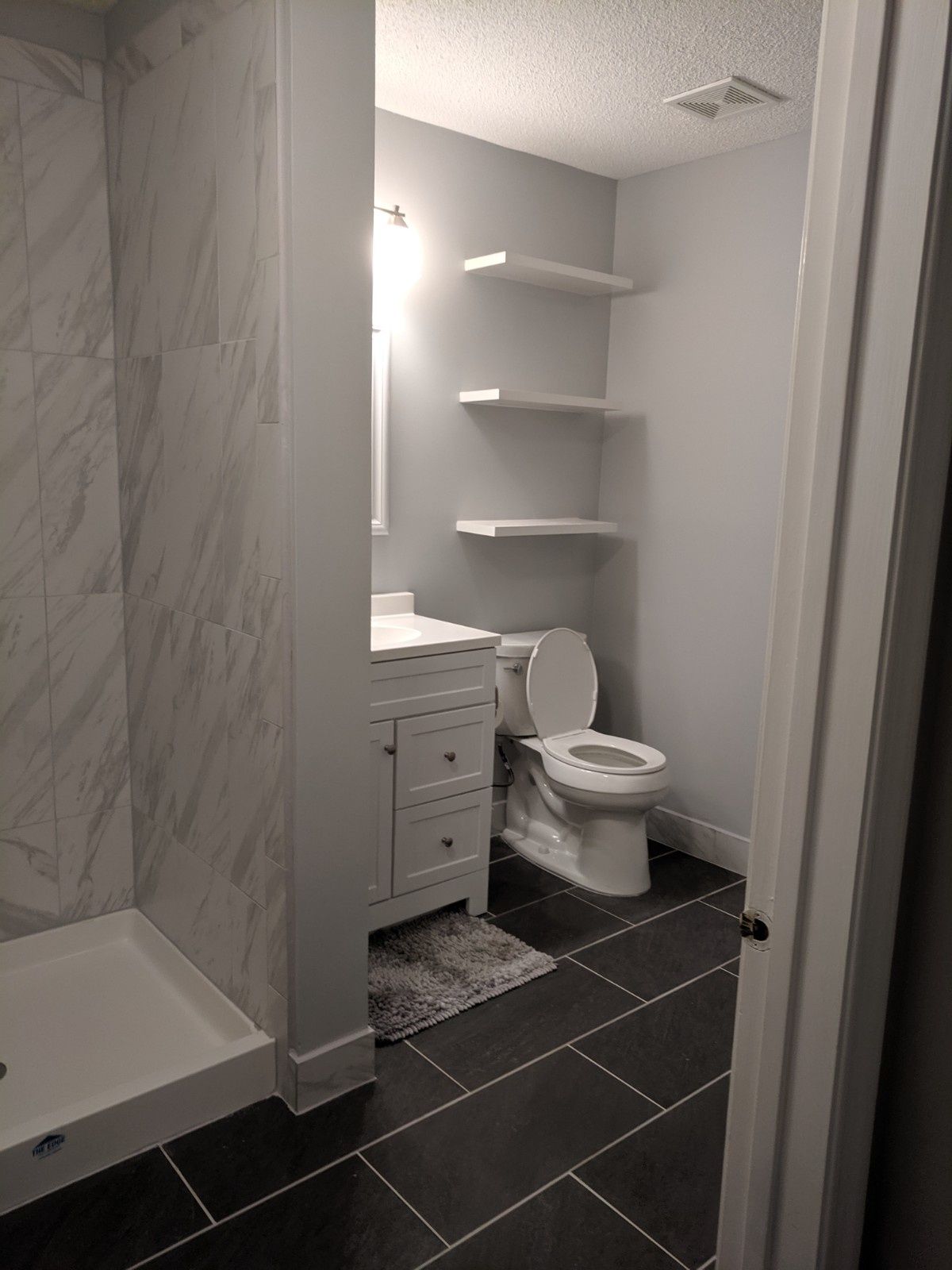 A modern bathroom with a white vanity, toilet, three floating wall shelves, and a gray-tiled walk-in shower.