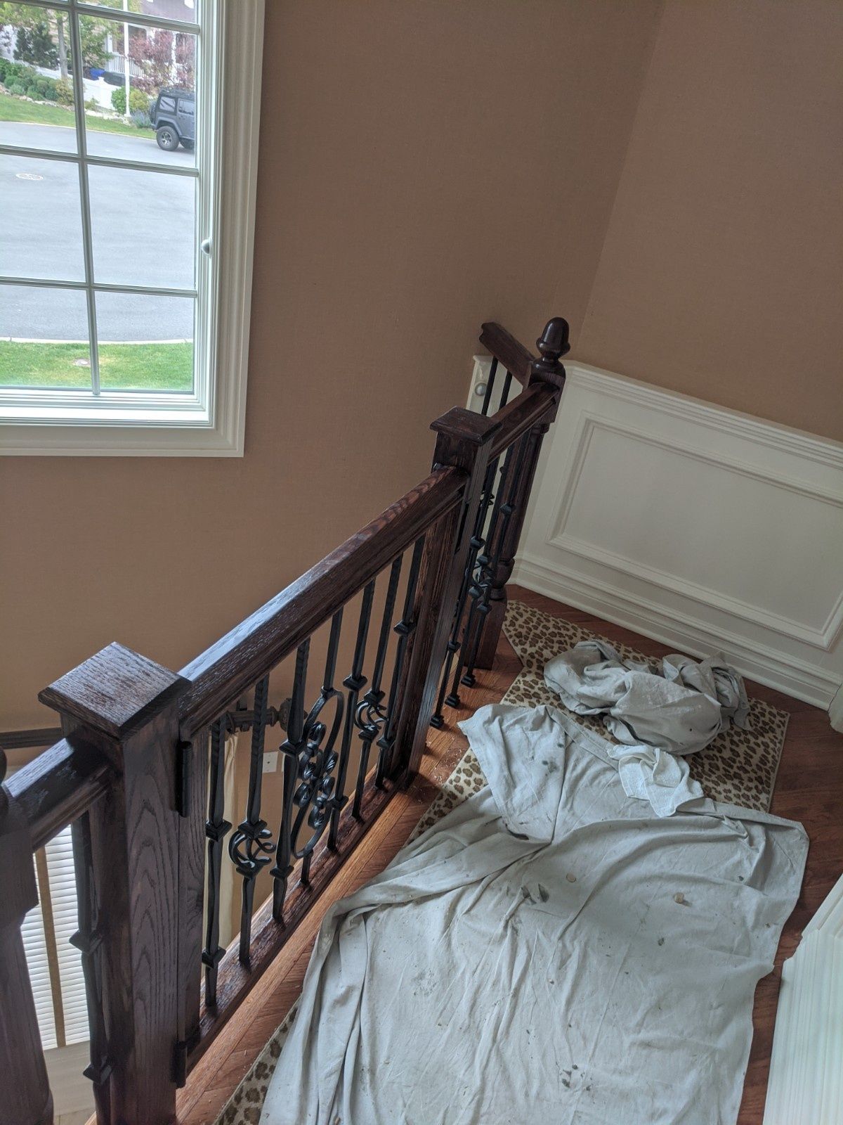 A dark wood stair railing with decorative metal balusters next to a window and a wall with white wainscoting and drop cloths.