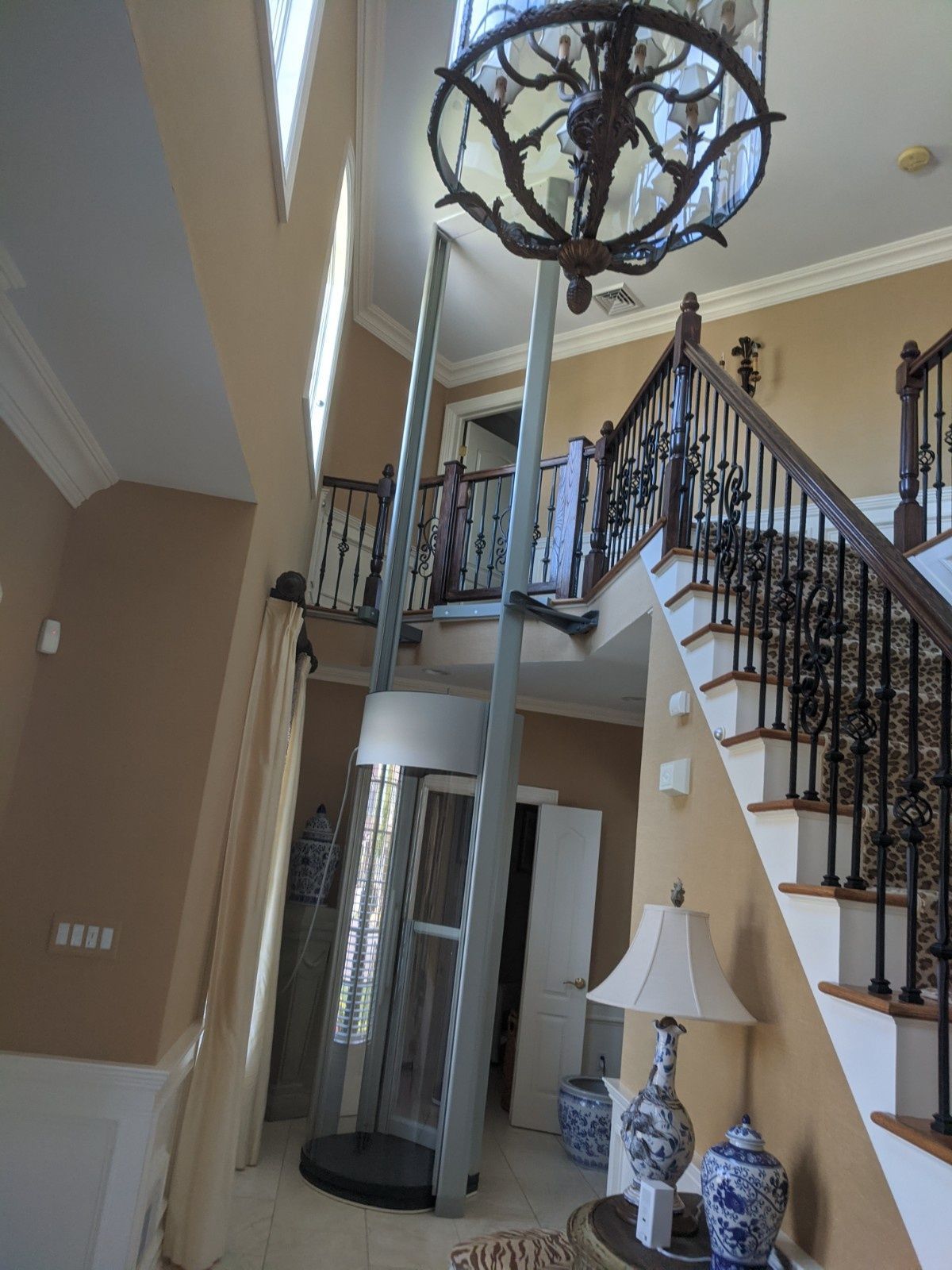 A tall, cylindrical residential glass elevator installed in the foyer of a home next to a wooden staircase.