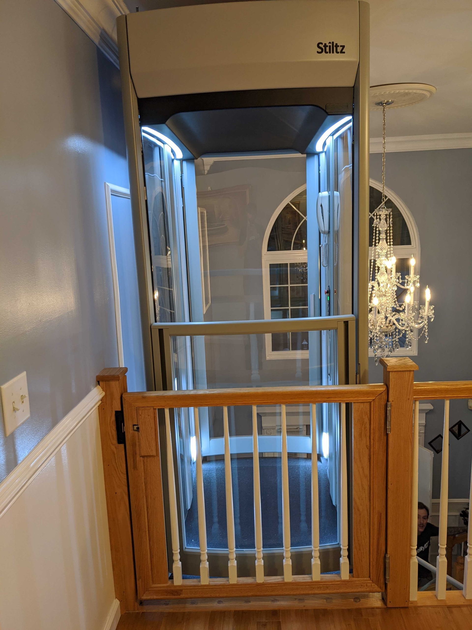 A residential glass elevator with white trim installed inside a home next to a wooden staircase railing.