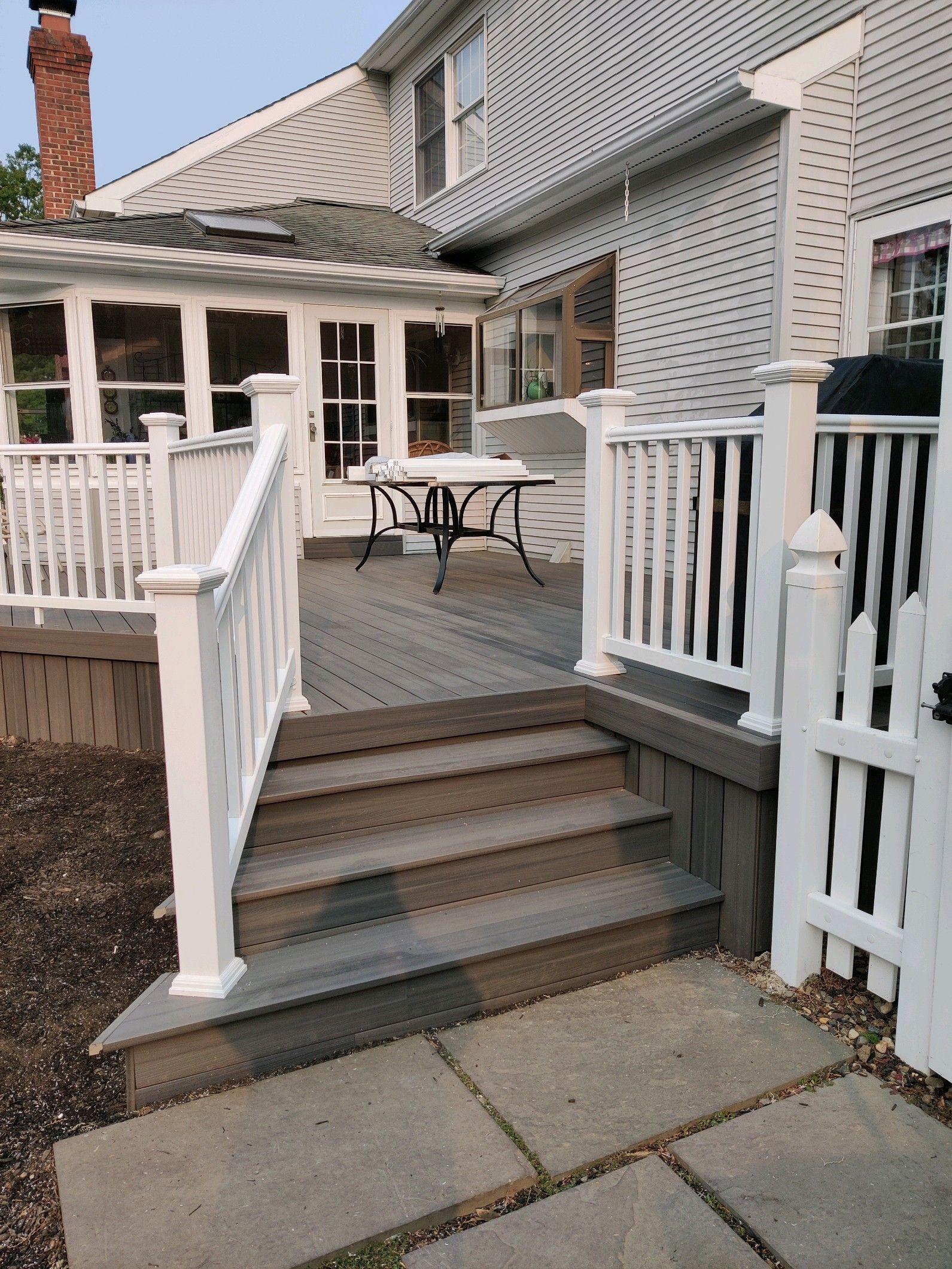 A wooden deck with white railings and stairs leading to a back patio with a glass-topped table.