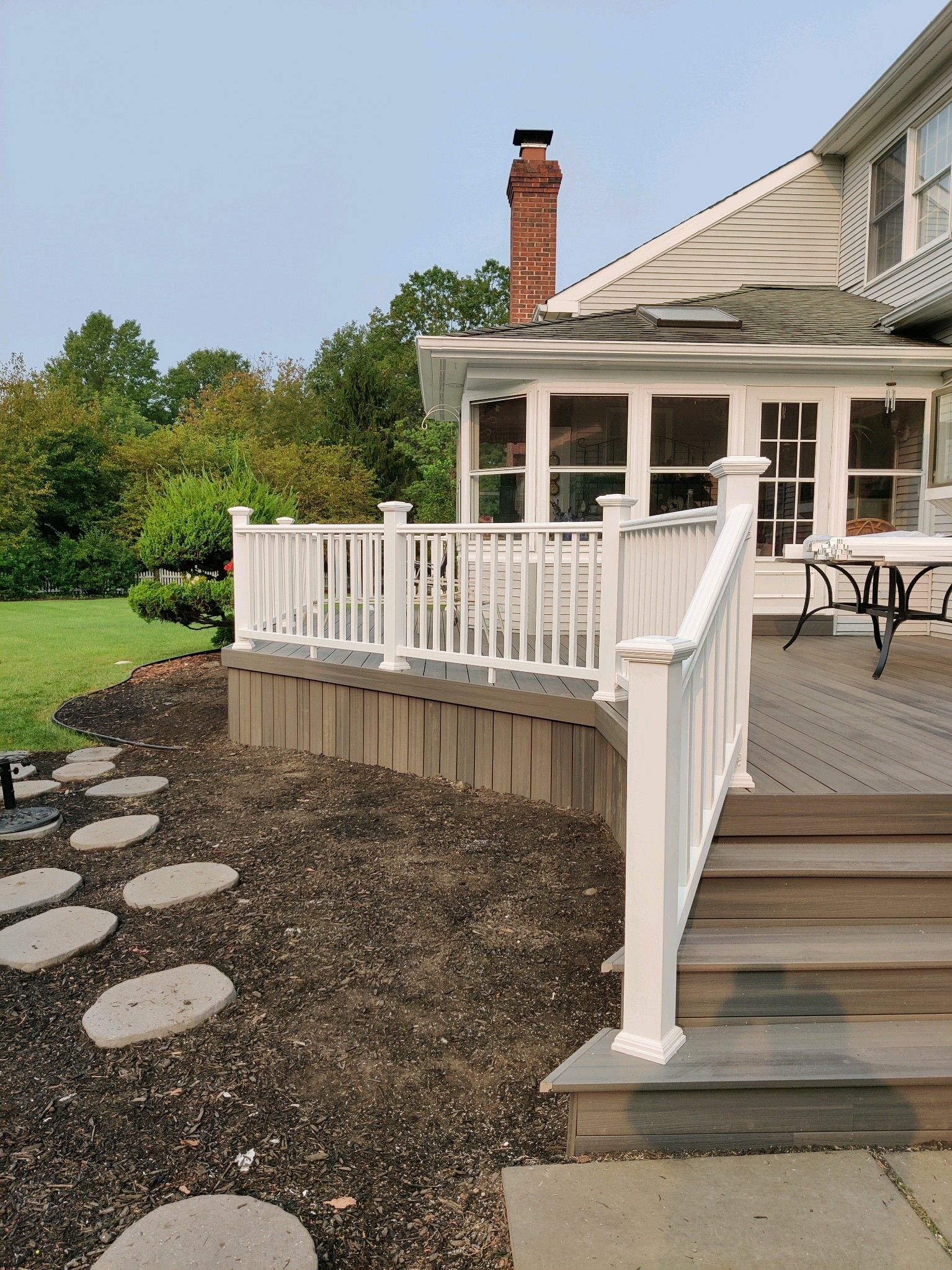 A wooden deck with white railings attached to a house with a sunroom, overlooking a lawn with stone stepping stones.