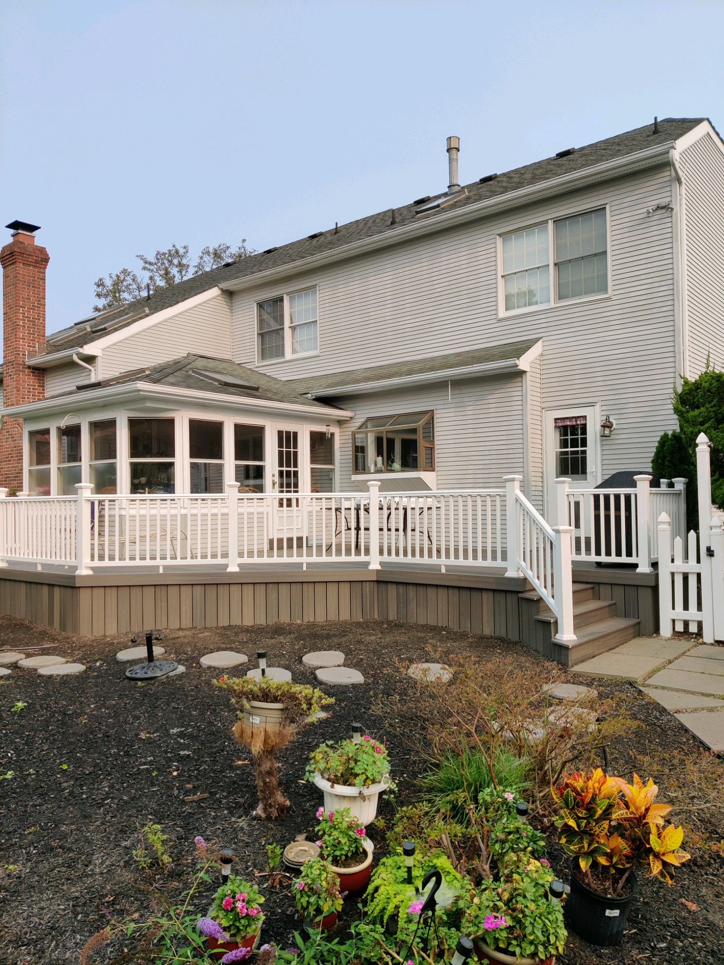 A two-story house with white siding and a wrap-around porch, featuring a gravel yard with potted plants in the foreground.