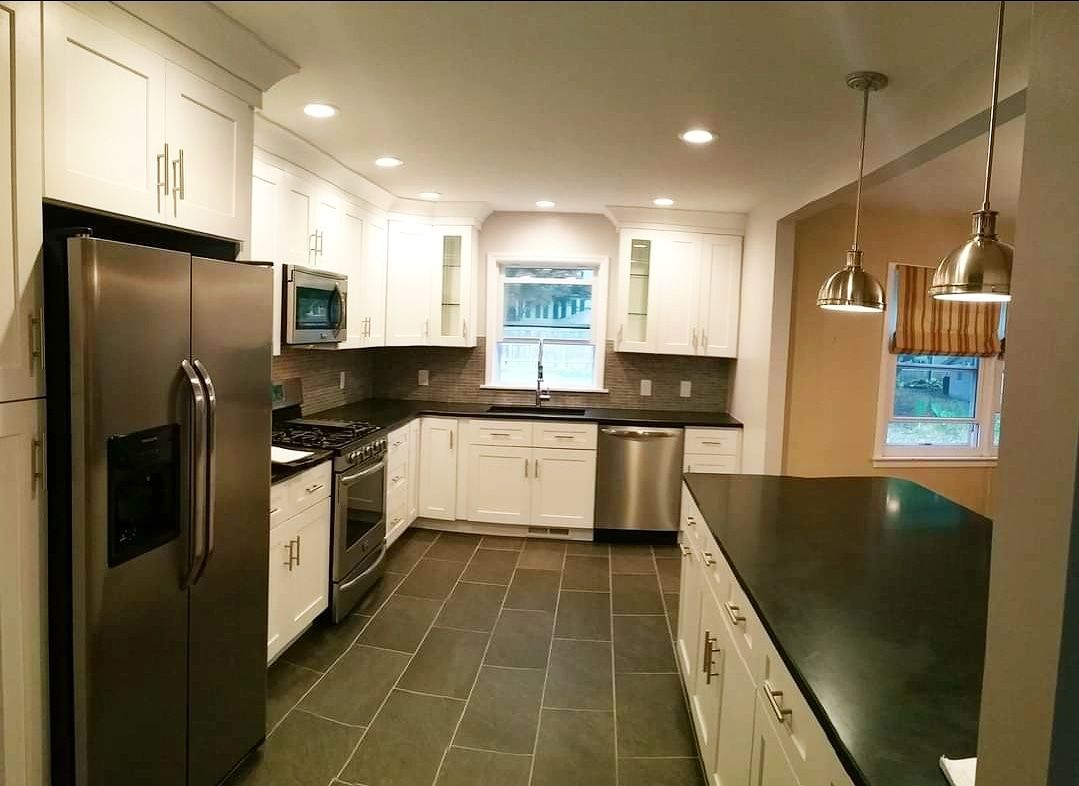 A modern kitchen with white cabinets, stainless steel appliances, dark countertops, and tile flooring under recessed lights.