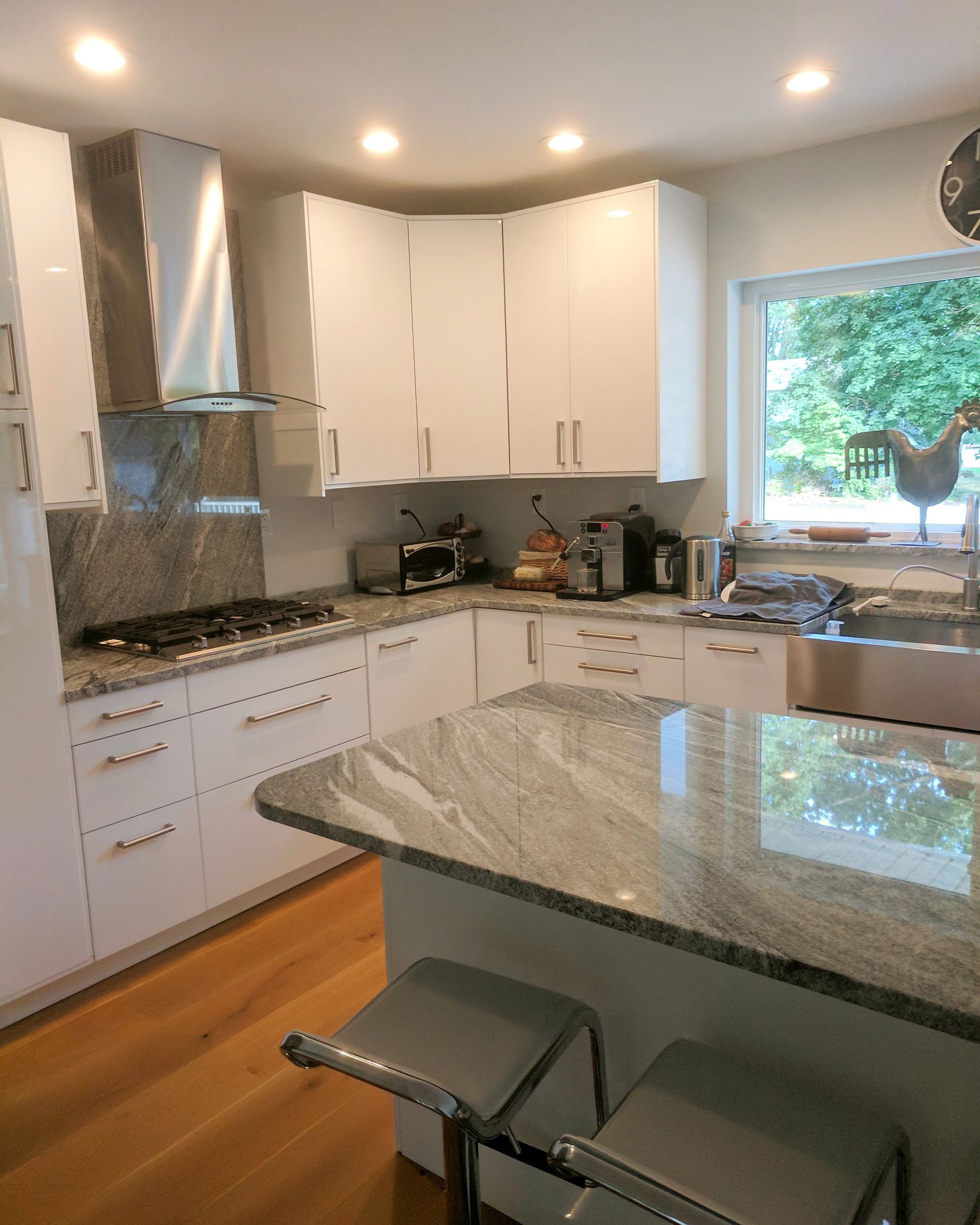 A modern kitchen with white cabinets, light wood flooring, gray granite countertops, and two stools at a kitchen island.