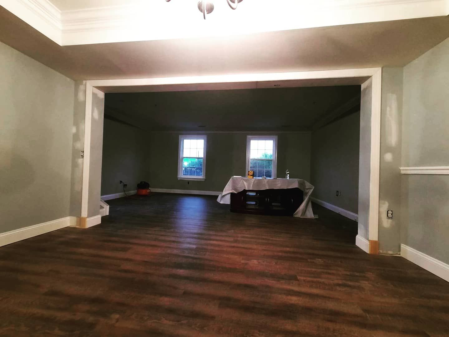 A wide, empty room with dark wood flooring, sage green walls under renovation, and a table covered in a white cloth.