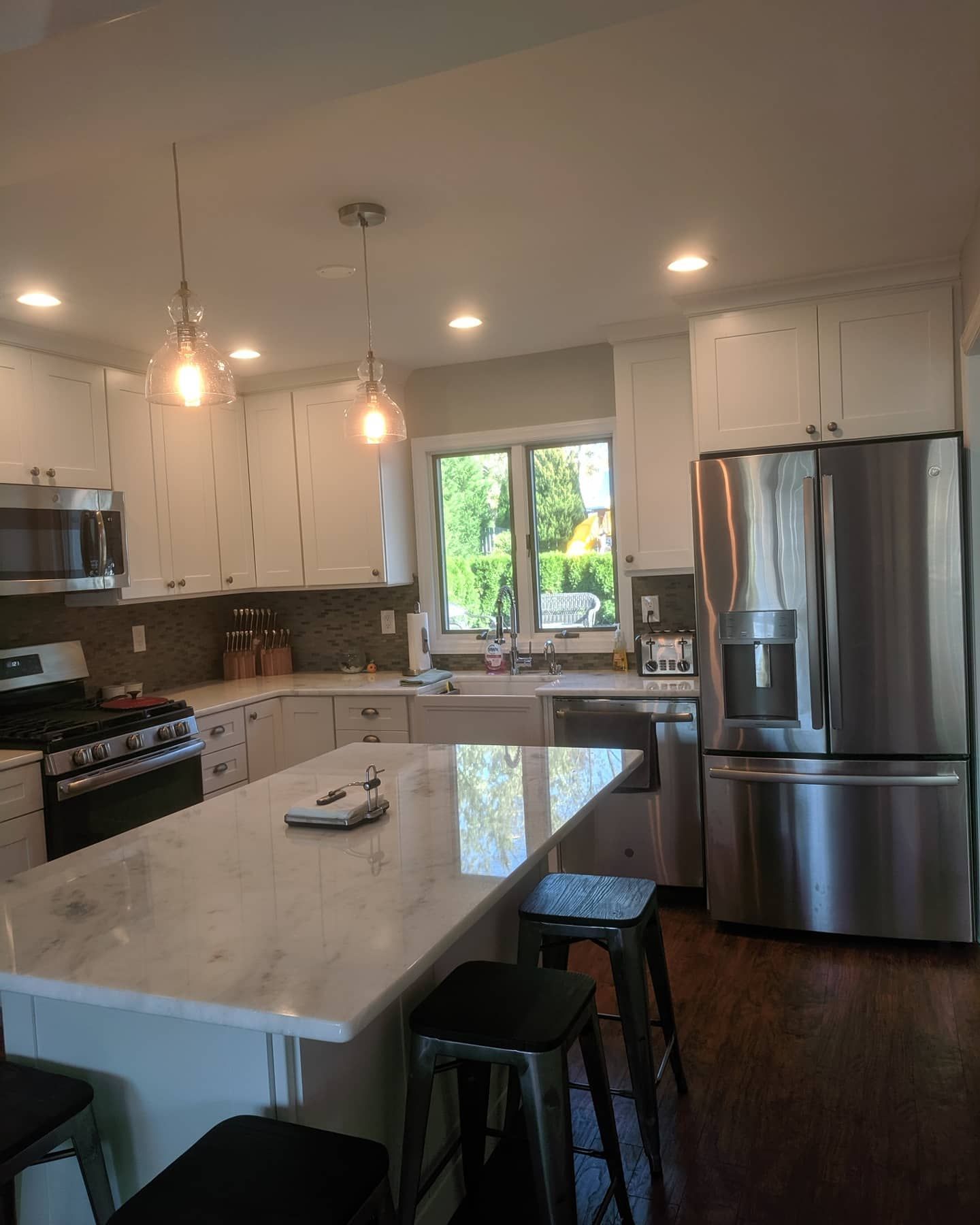 A modern kitchen featuring white cabinets, a marble-topped island with stools, stainless appliances, and a window.