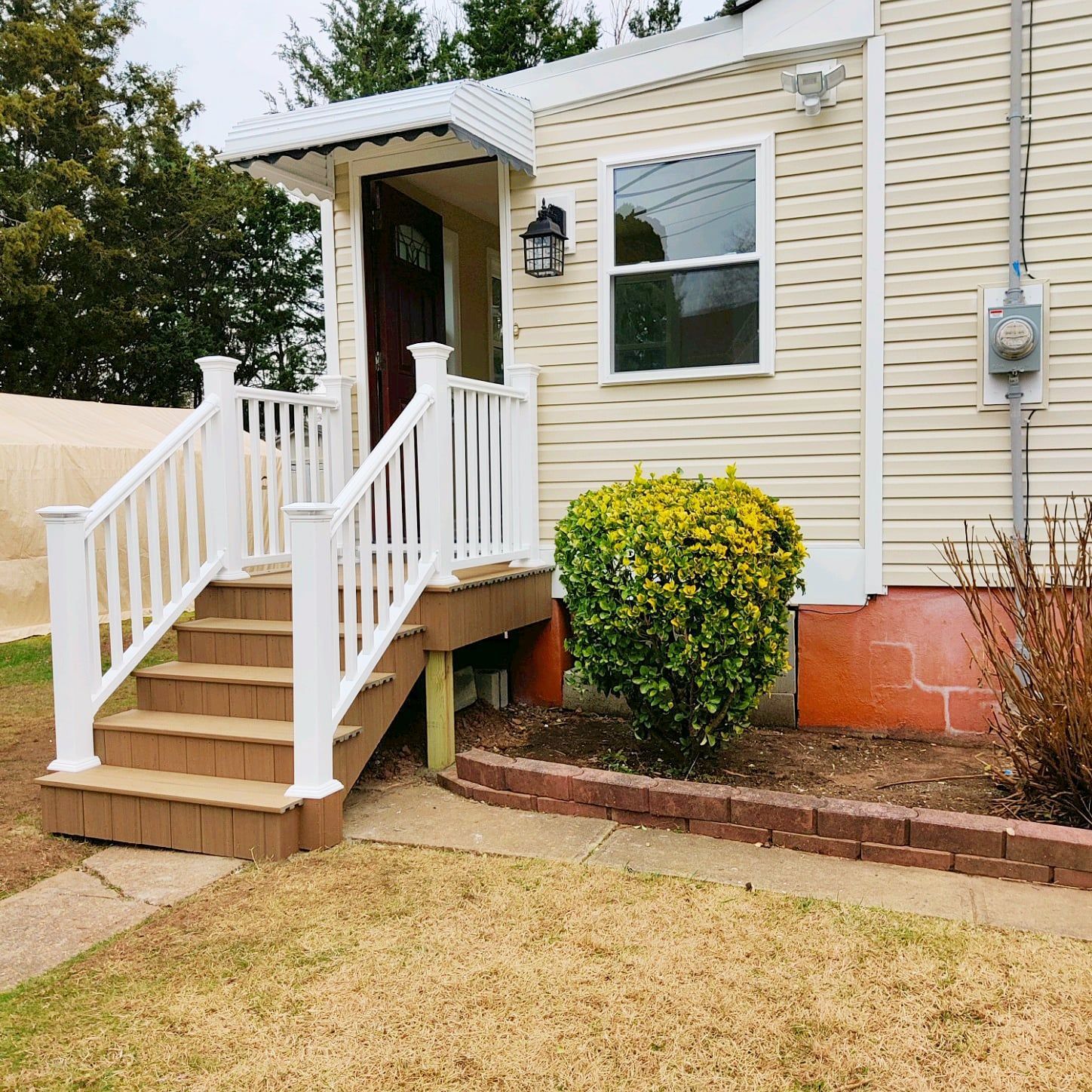A beige house with white trim, a small wooden staircase with white railings, a front door, and a green shrub.
