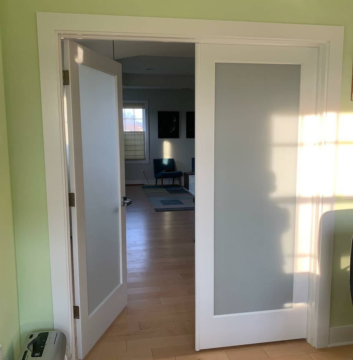 A pair of white, frosted glass French doors standing partially open, revealing a bright room with hardwood floors.
