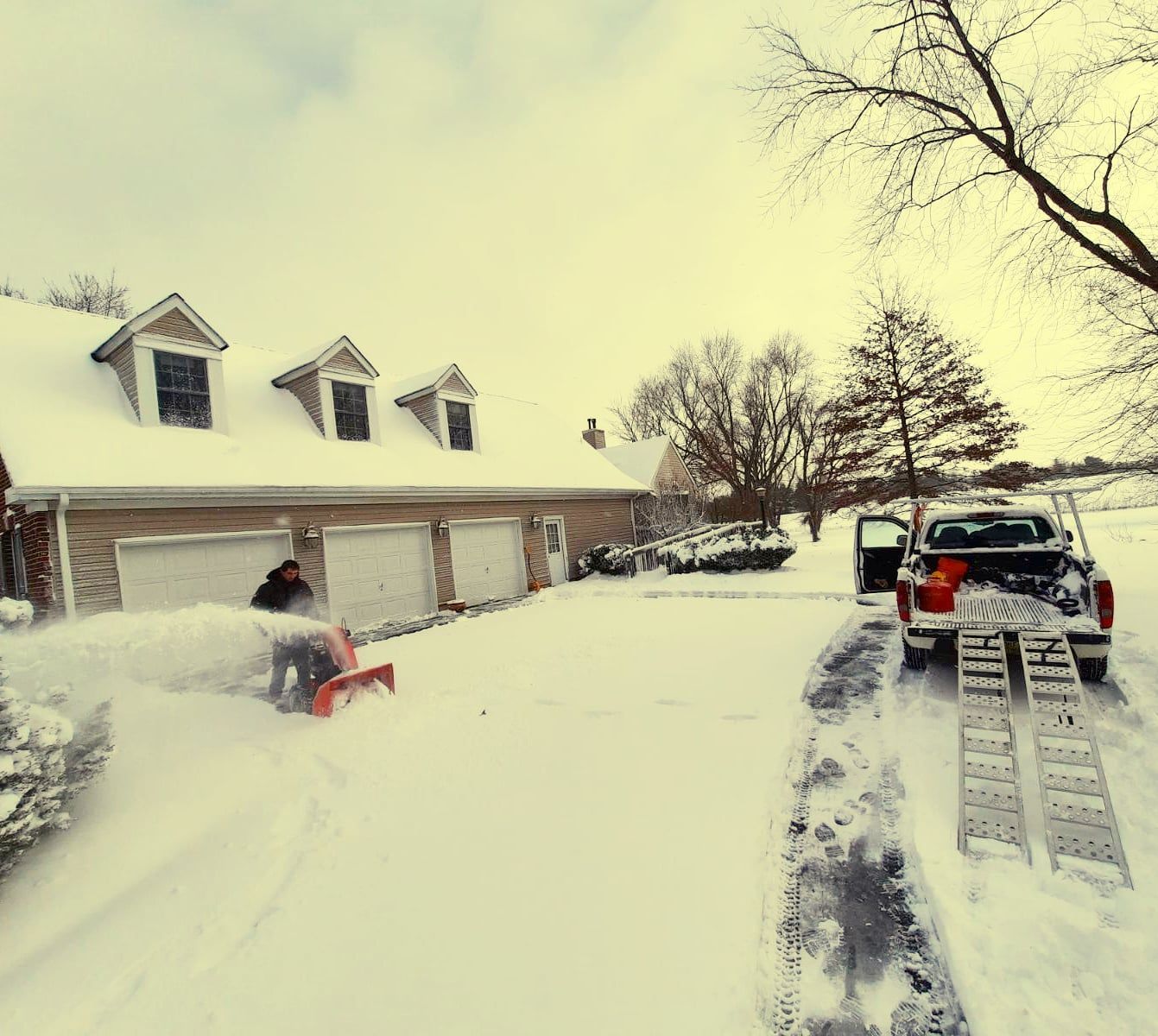 A person uses a red snowblower to clear a snow-covered driveway in front of a three-car garage, near a pickup truck.