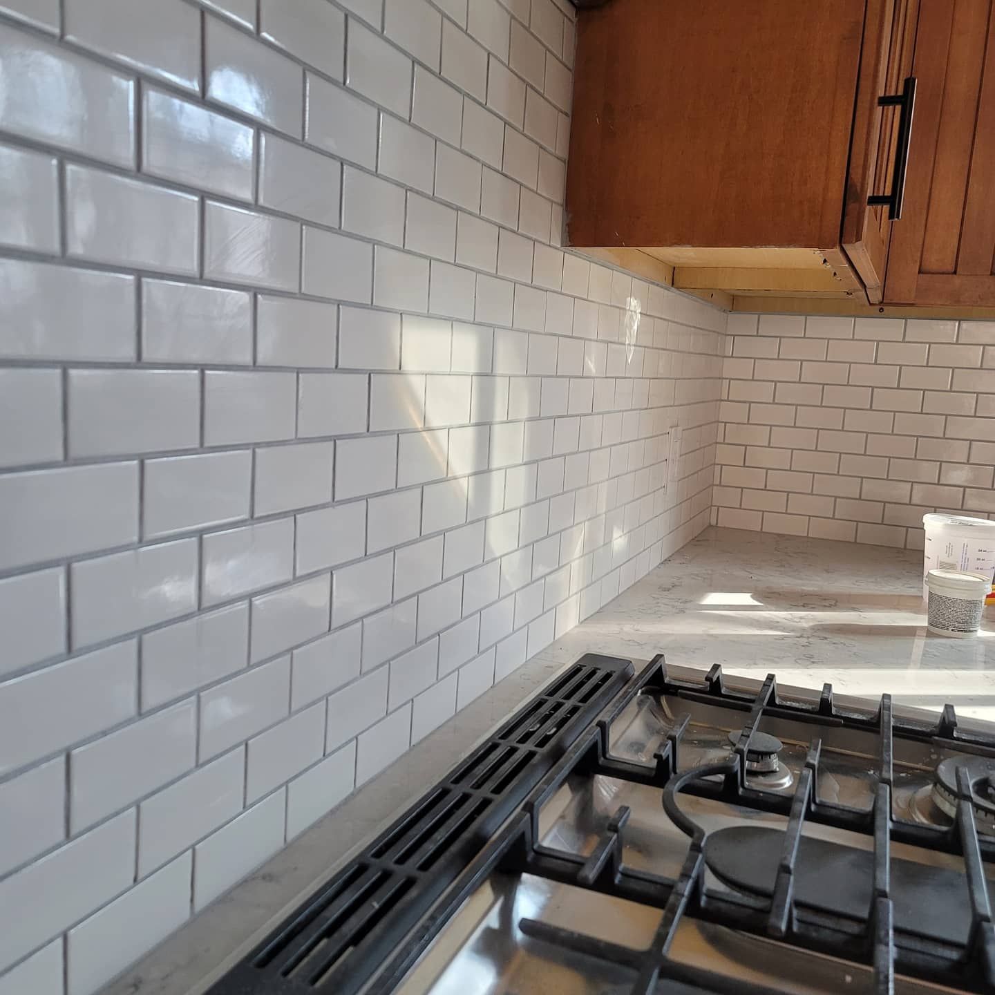 A white subway tile backsplash installed behind a stainless steel gas stovetop and under wooden kitchen cabinets.