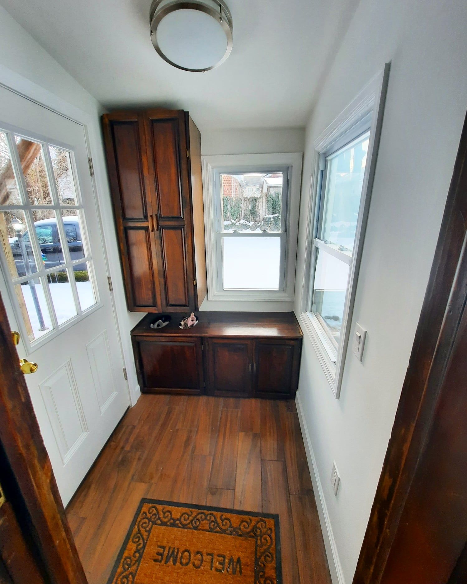 An entryway with a white door, wood-look flooring, built-in dark wood cabinets, a bench, and two windows.
