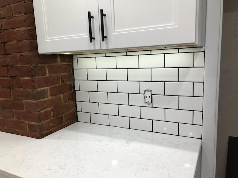 White subway tile backsplash with dark grout, installed under white cabinets next to a red brick wall and marble counter.