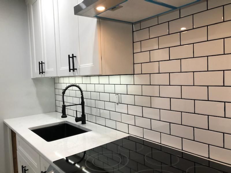 A kitchen counter with a black sink, faucet, and stovetop against a white subway tile backsplash and white cabinets.