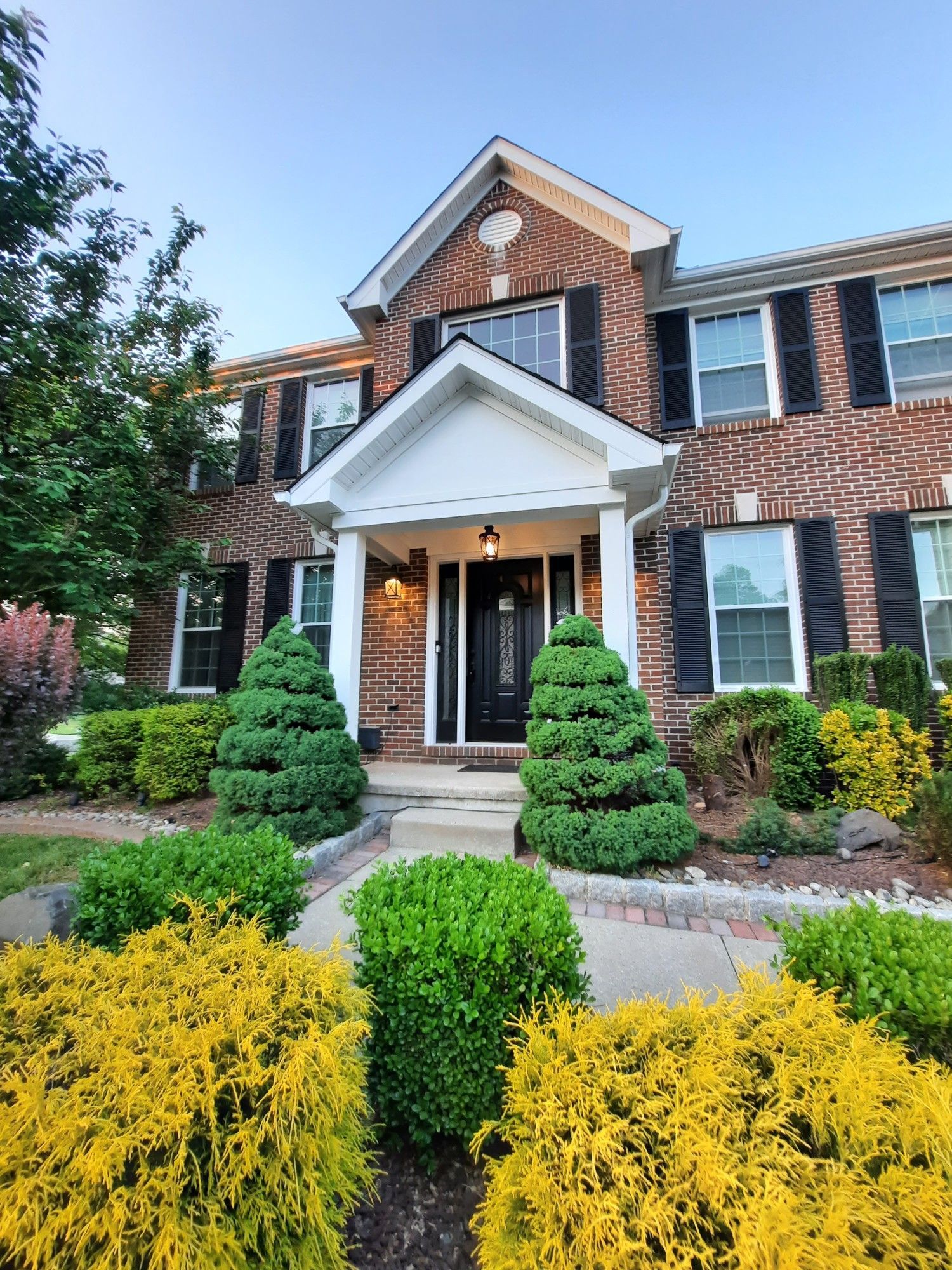 A brick suburban home with a white covered entryway, surrounded by green and yellow manicured shrubs under a blue sky.
