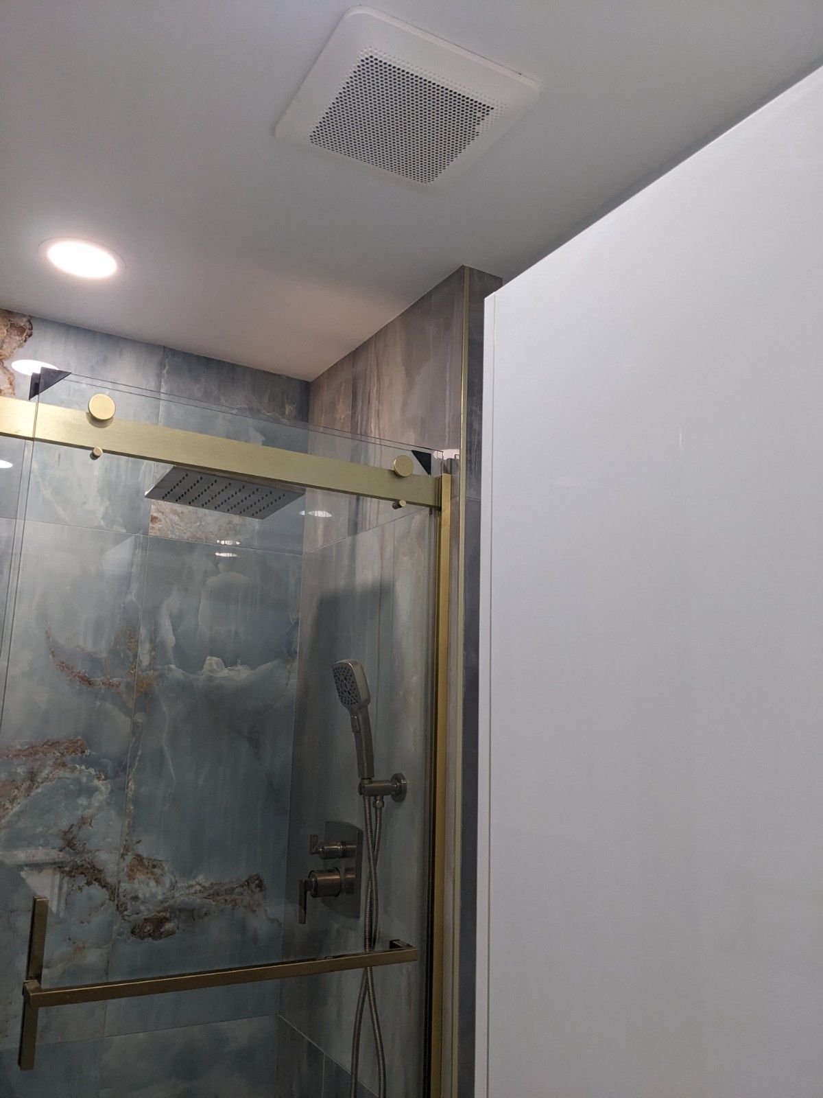 A gold-trimmed glass shower enclosure with stone-patterned tiles, a handheld showerhead, and a ceiling ventilation fan.