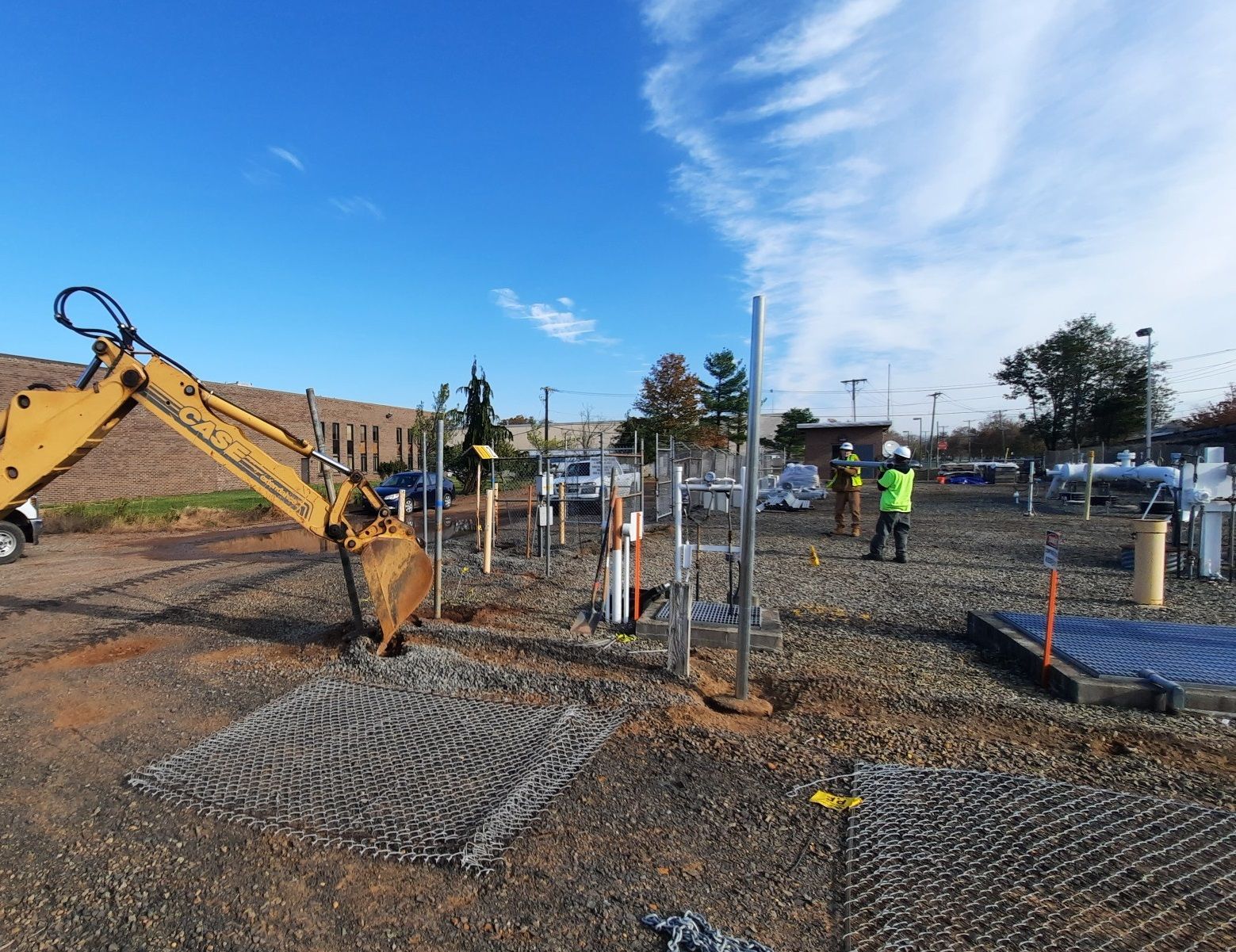 Construction workers near a yellow excavator and metal pipes at a graveled, open-air work site under a blue sky.