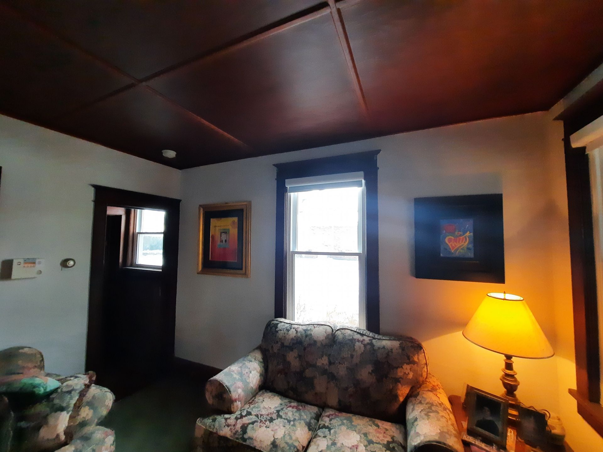 A floral loveseat sits below a window in a cozy living room with a dark paneled ceiling and soft lamp lighting.