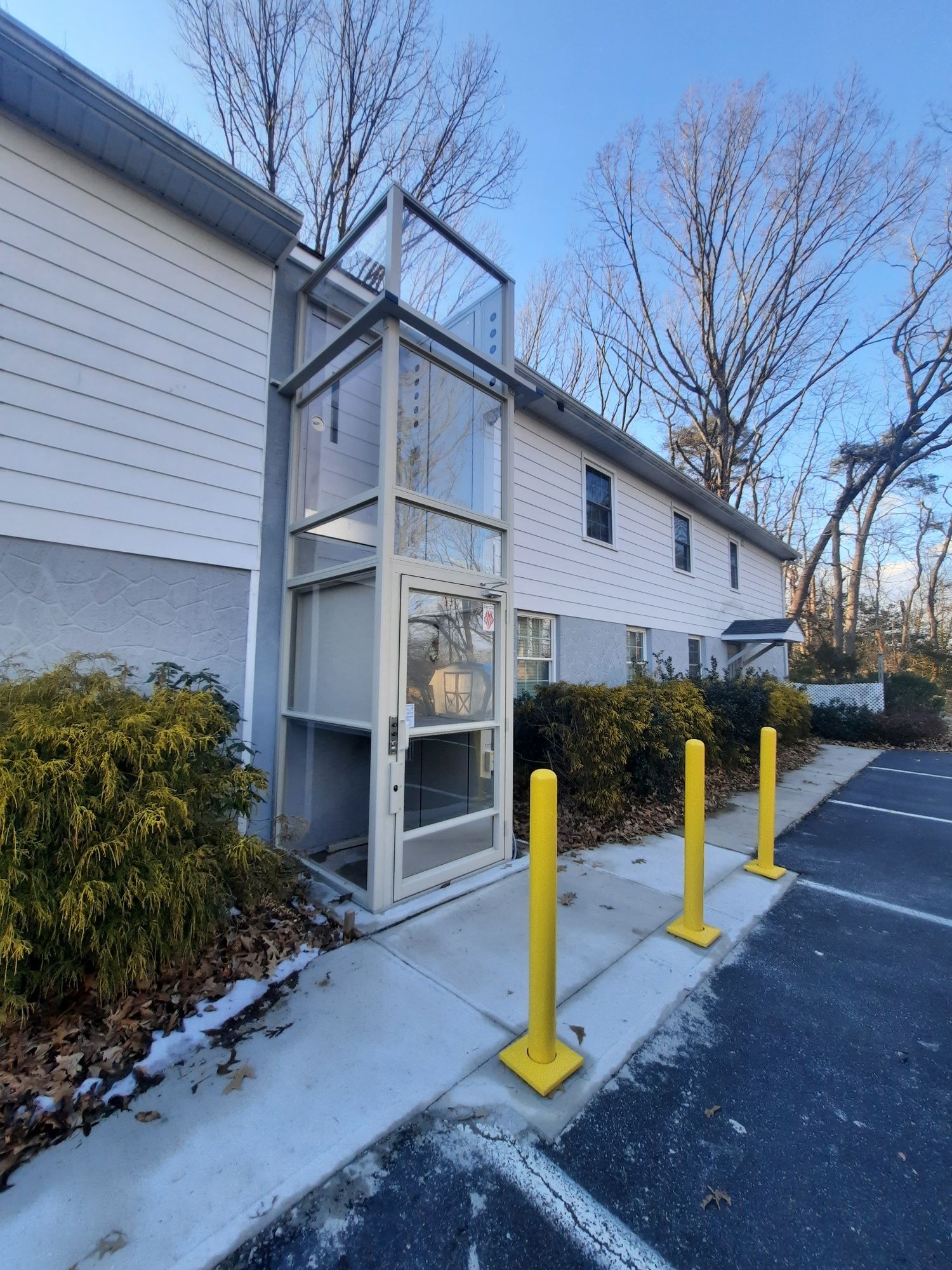 An exterior vertical platform lift for accessibility attached to a multi-story building, with yellow safety bollards.