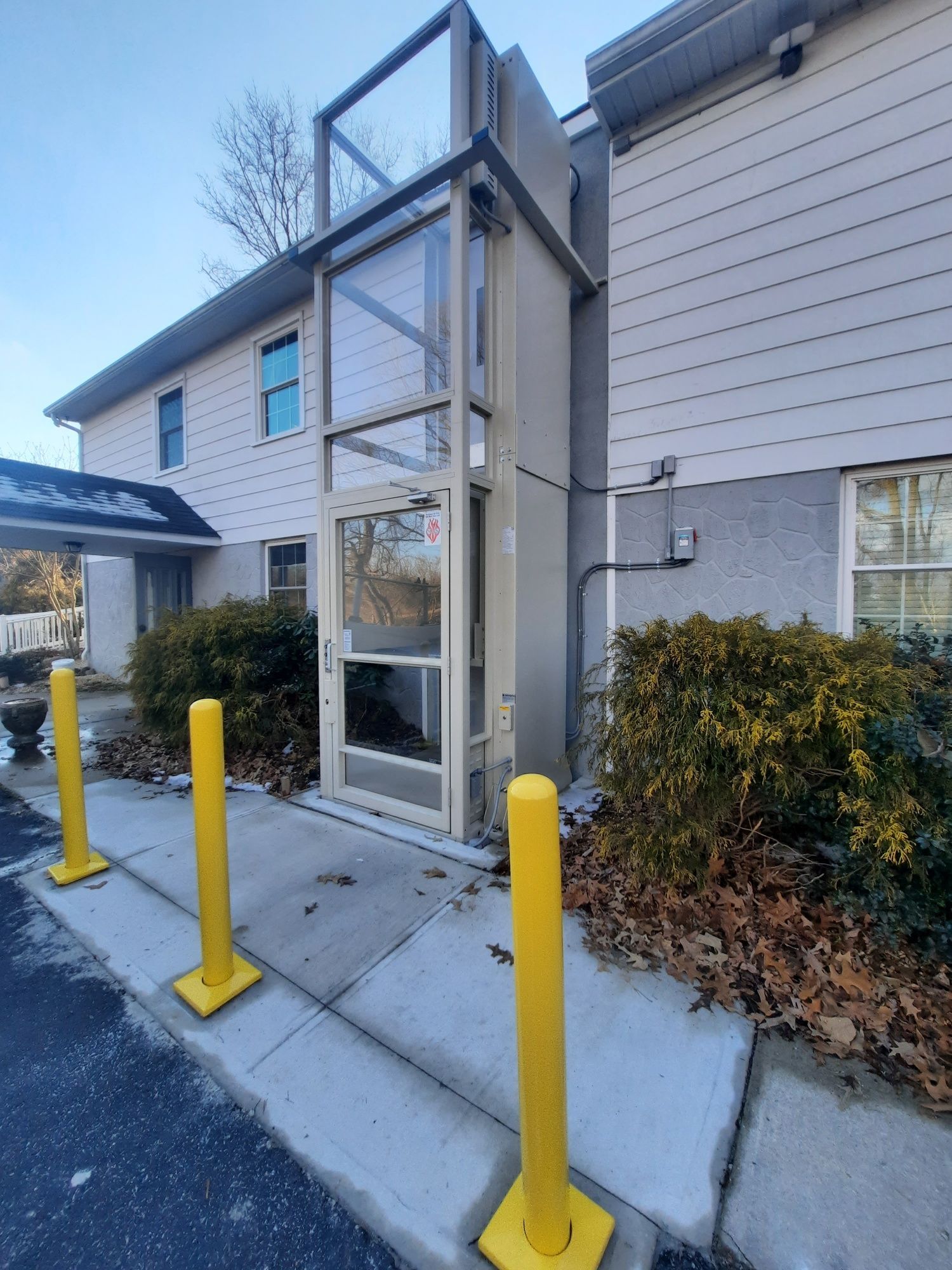 An exterior vertical wheelchair lift with a glass enclosure stands against a two-story building near yellow safety bollards.