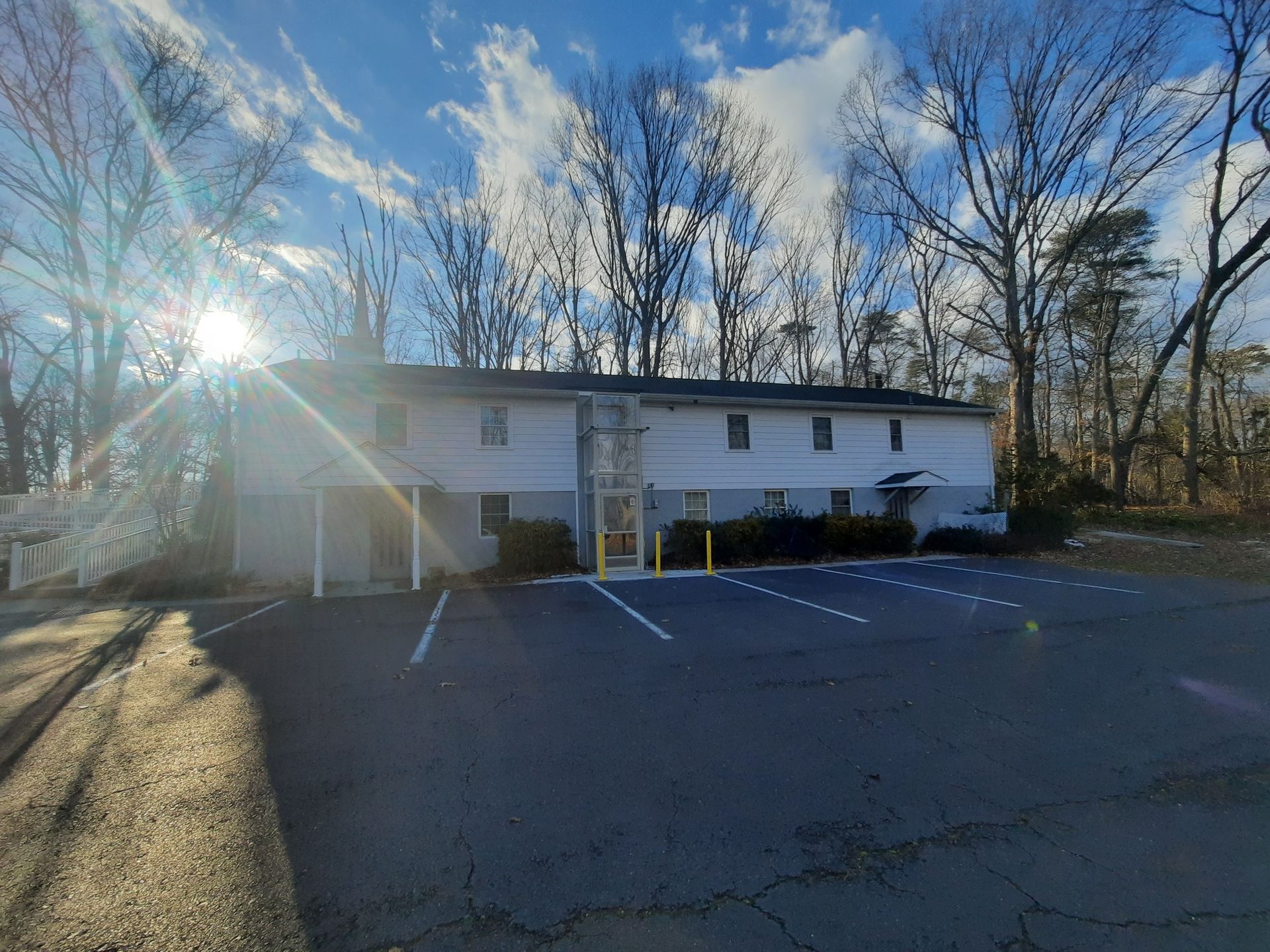 Two-story office building with white siding, grey ground level, and a paved parking lot under a sunny blue sky.
