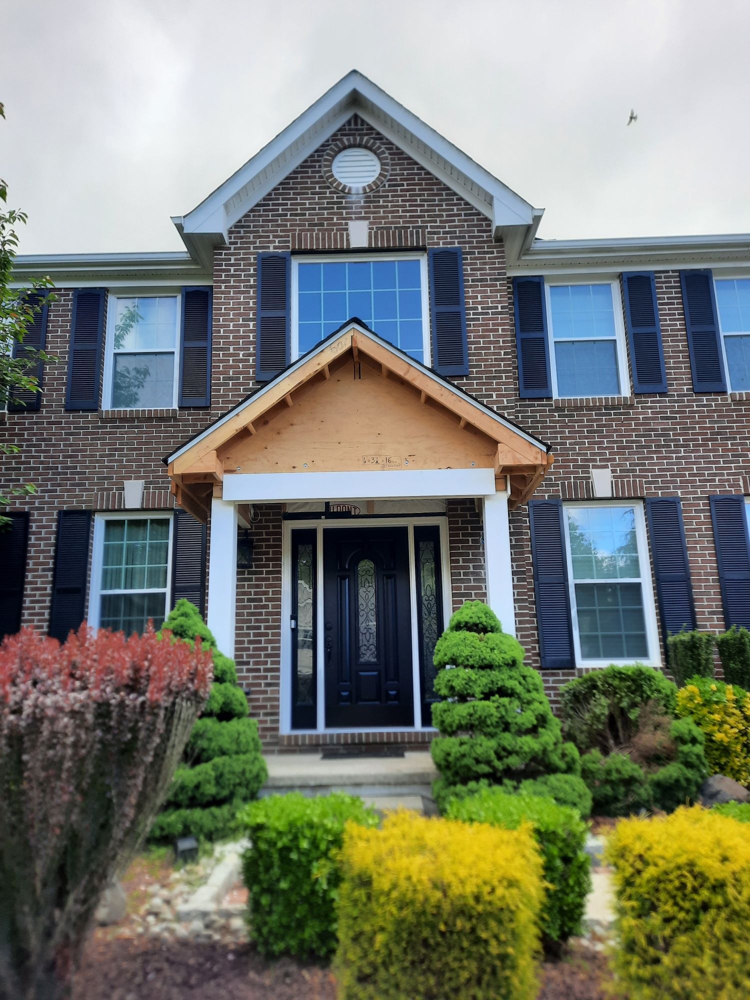 Two-story brick house with a front door under a wooden-framed portico, flanked by dark shutters and manicured shrubs.
