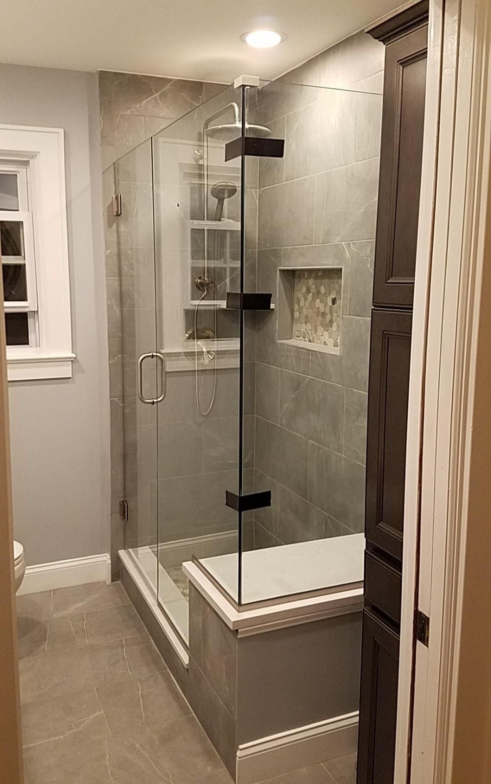 A modern tiled walk-in shower with a glass door, a built-in bench, and a niche in a bathroom with grey flooring.