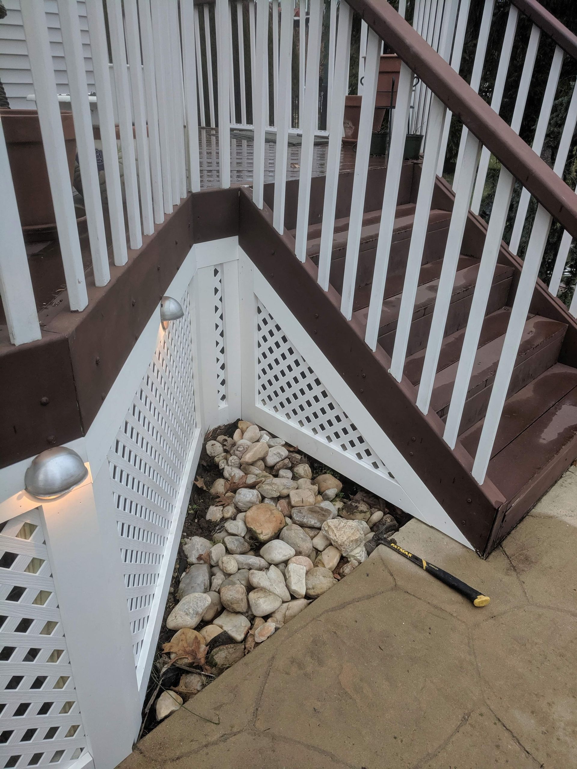 A corner of a wooden deck staircase with white lattice skirting and a ground-level rock garden bed on a stone patio.