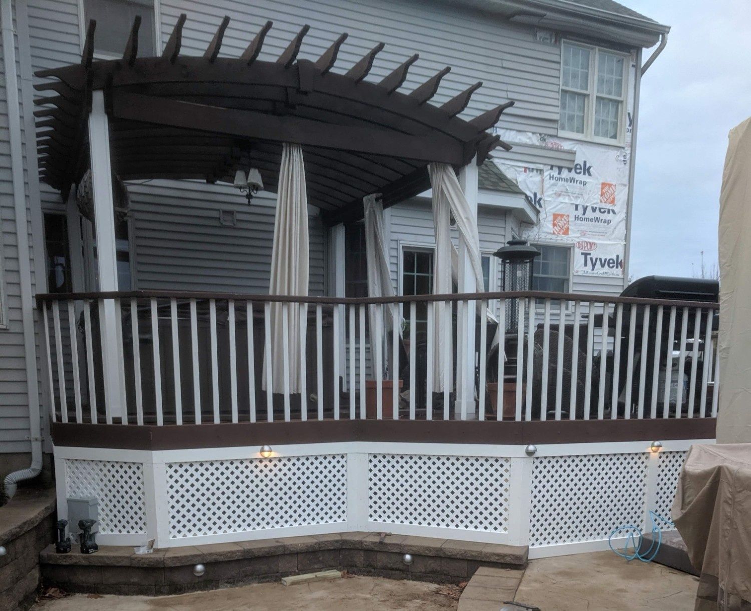 A deck with a curved dark brown pergola, white railings, and white lattice skirting on the side of a house.