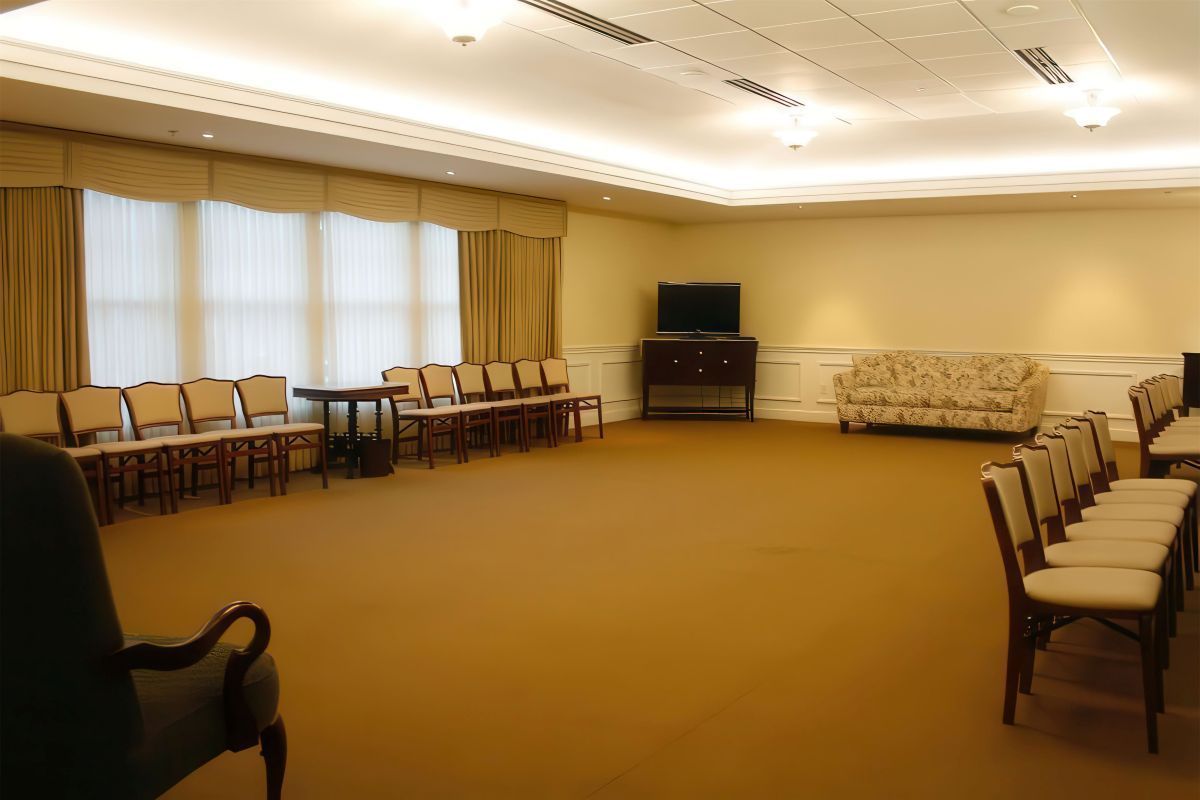 Empty room with rows of chairs, windows, and sofa; beige walls and carpet.