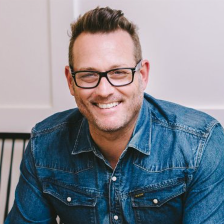 A man wearing glasses and a denim shirt smiles for the camera