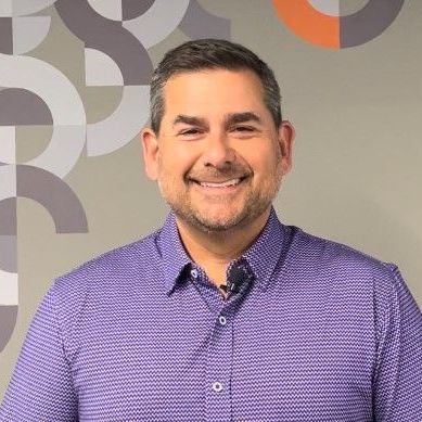 A man in a purple shirt is smiling in front of a wall with geometric shapes.