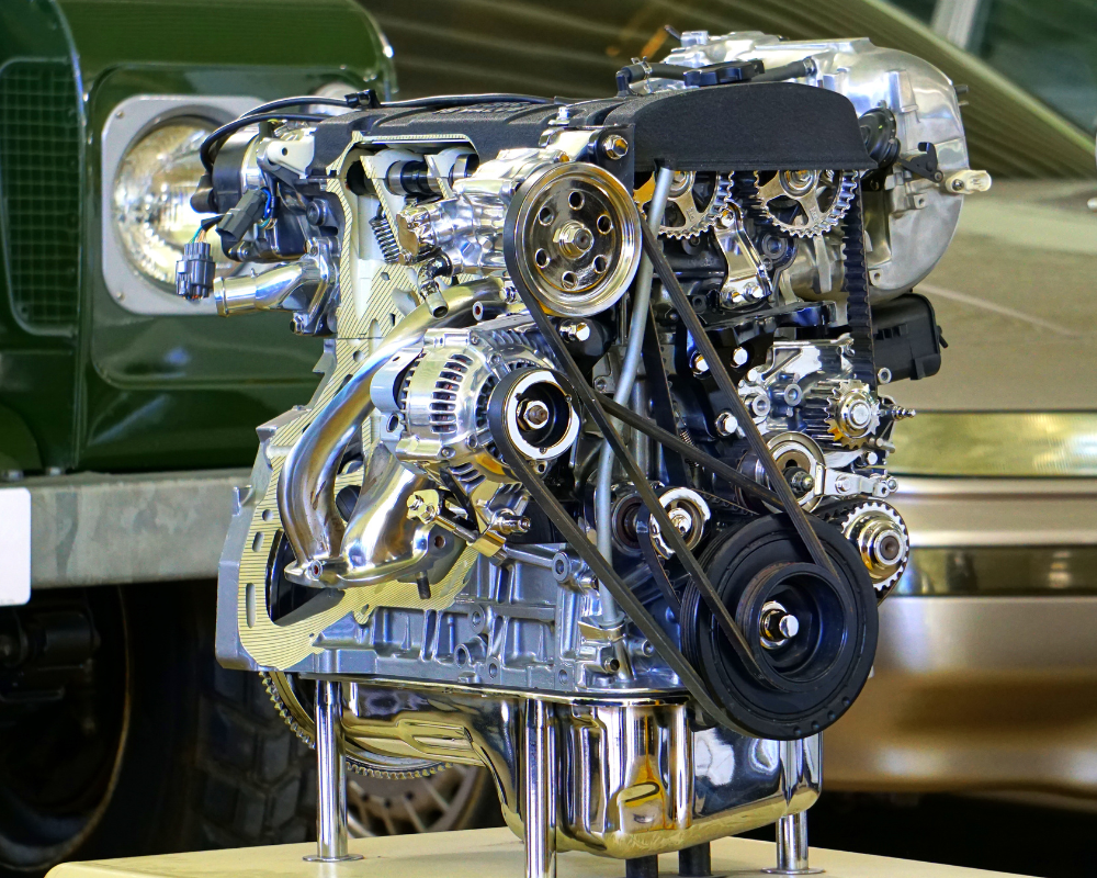 Shiny engine with belts and pulleys, displayed on a stand. Partly obscured green vehicle in the background.