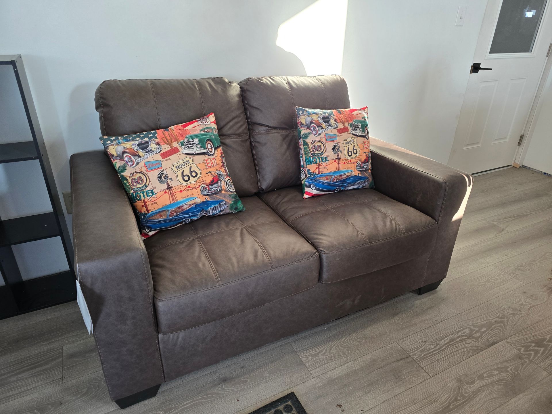 Brown loveseat with two colorful patterned pillows in a bright room with wood flooring and a white door