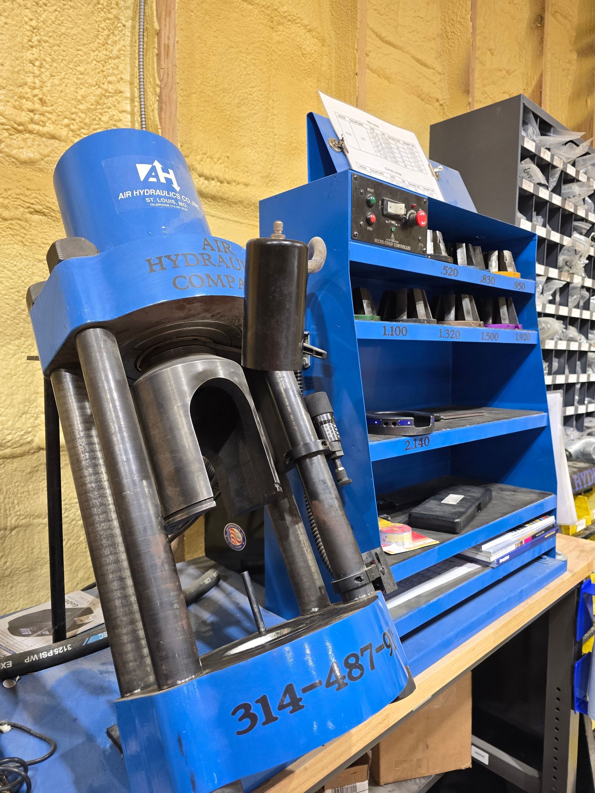 Blue industrial machine with metal rollers and a nearby blue shelving unit in a workshop.