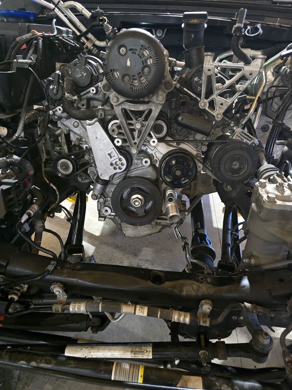 Person working on car engine, another person reviewing paperwork.