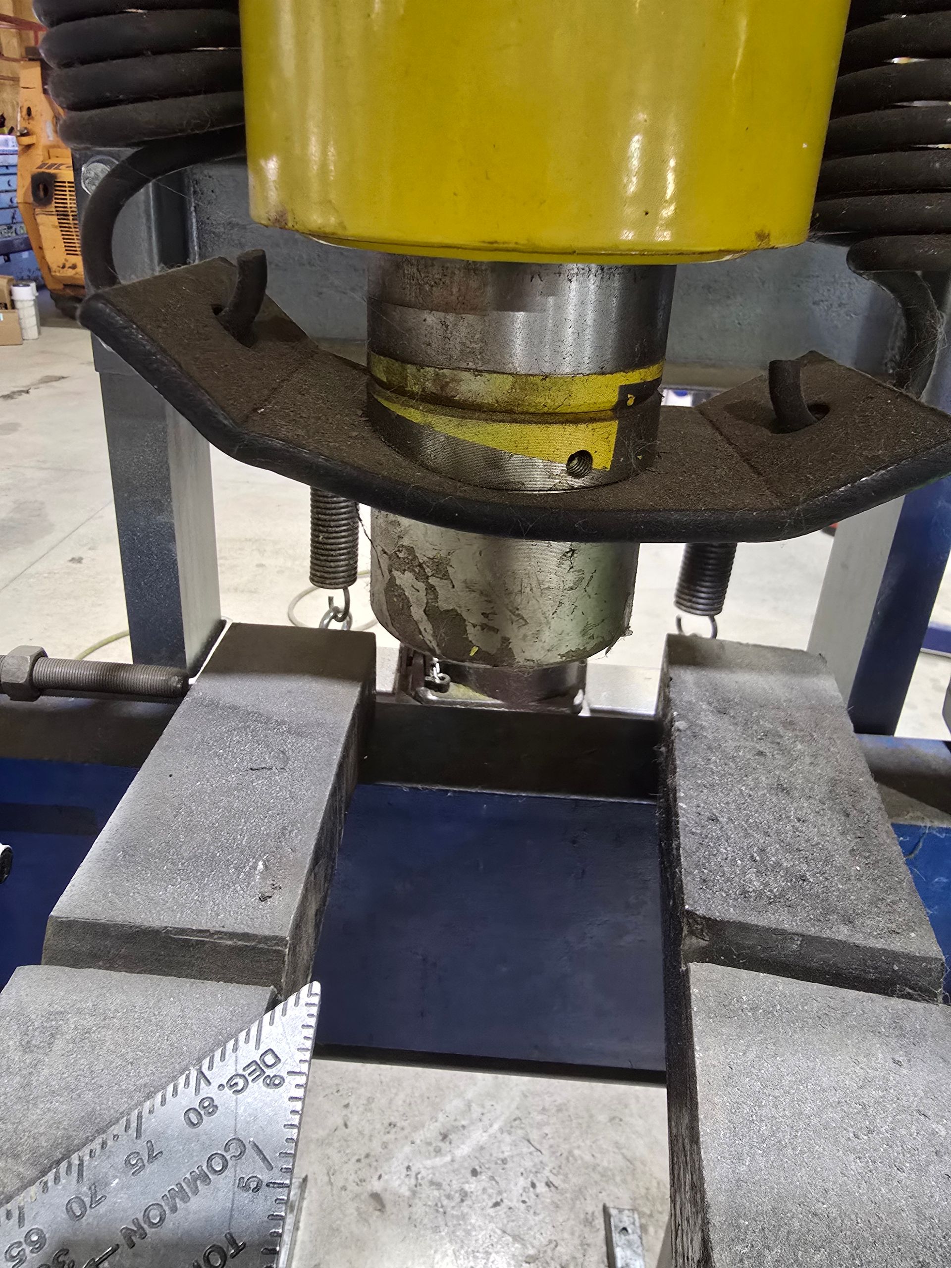 Close-up of a yellow hydraulic press compressing a dark block between two gray metal supports.
