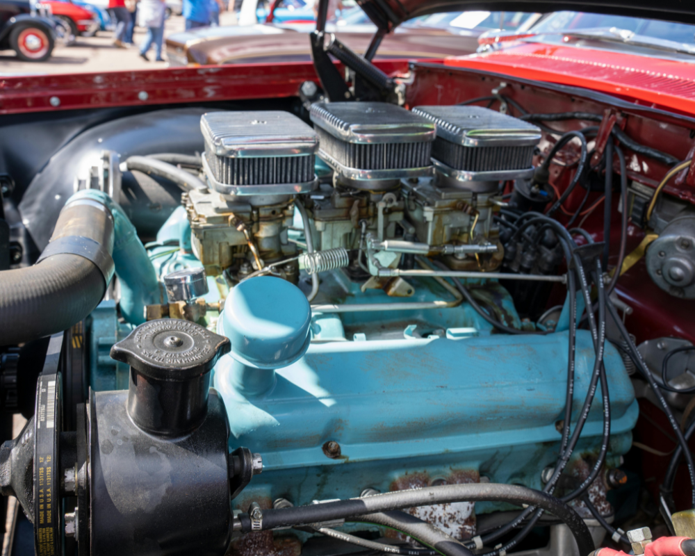 Classic red car engine with turquoise block and triple carburetors, outdoors.