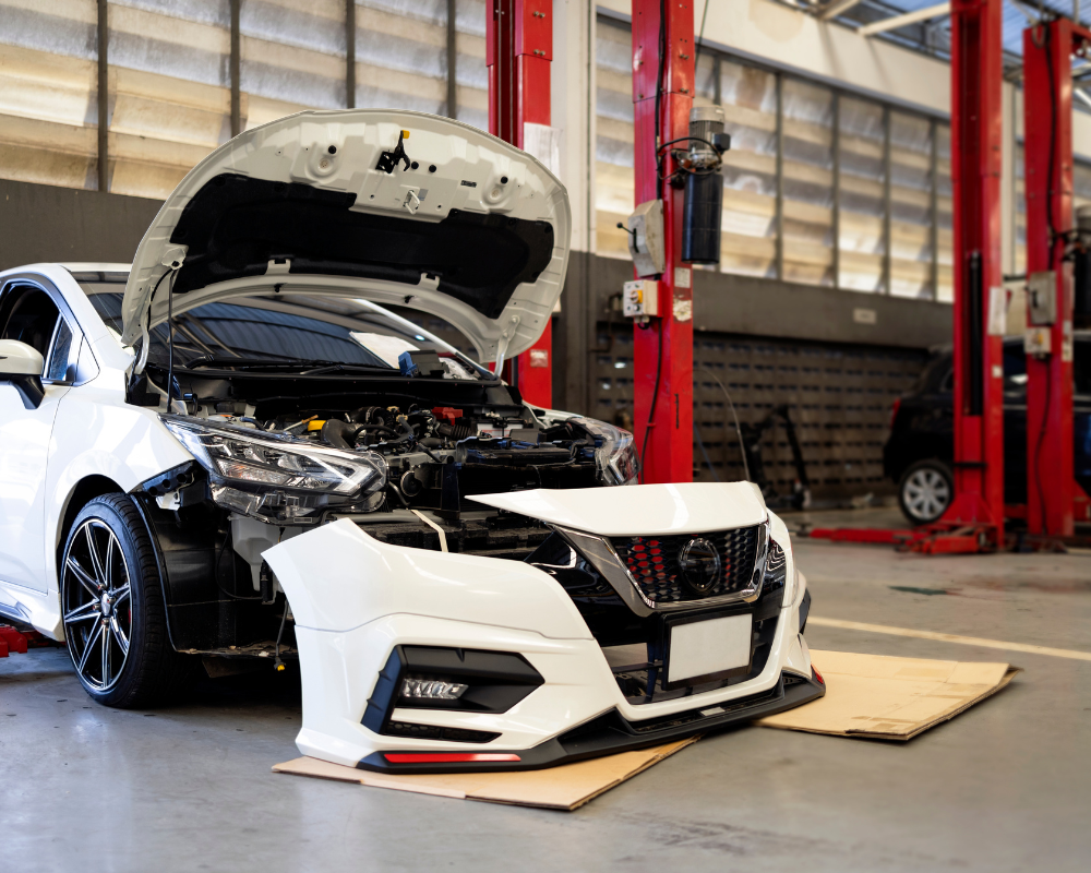 White car with open hood in a repair shop, front bumper detached, red lift columns.