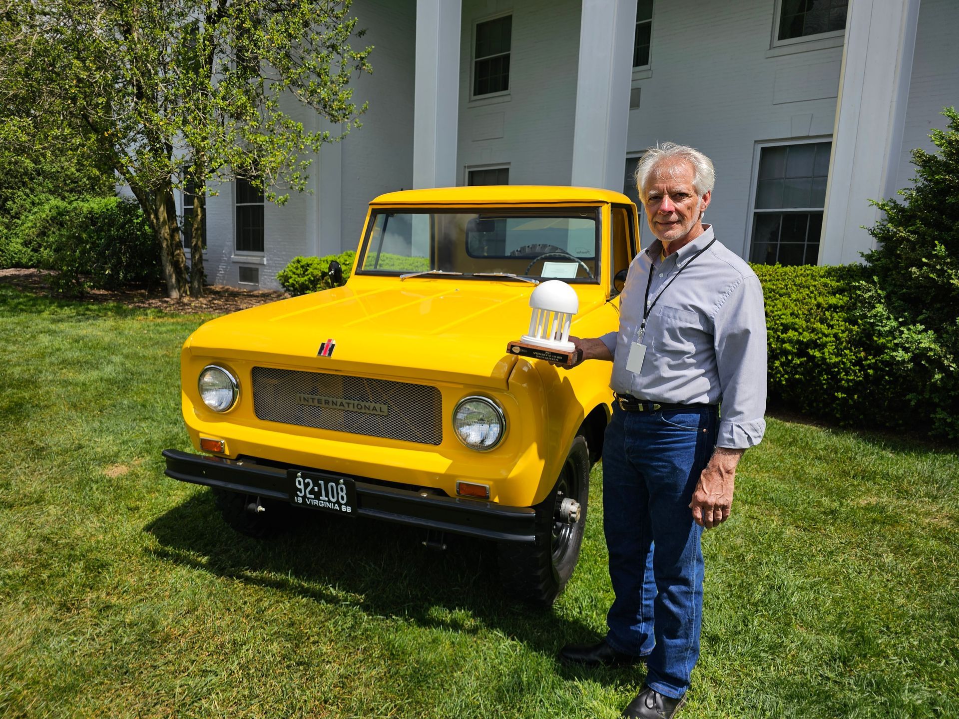 Man holding object stands beside a bright yellow vintage truck in front of a white building.