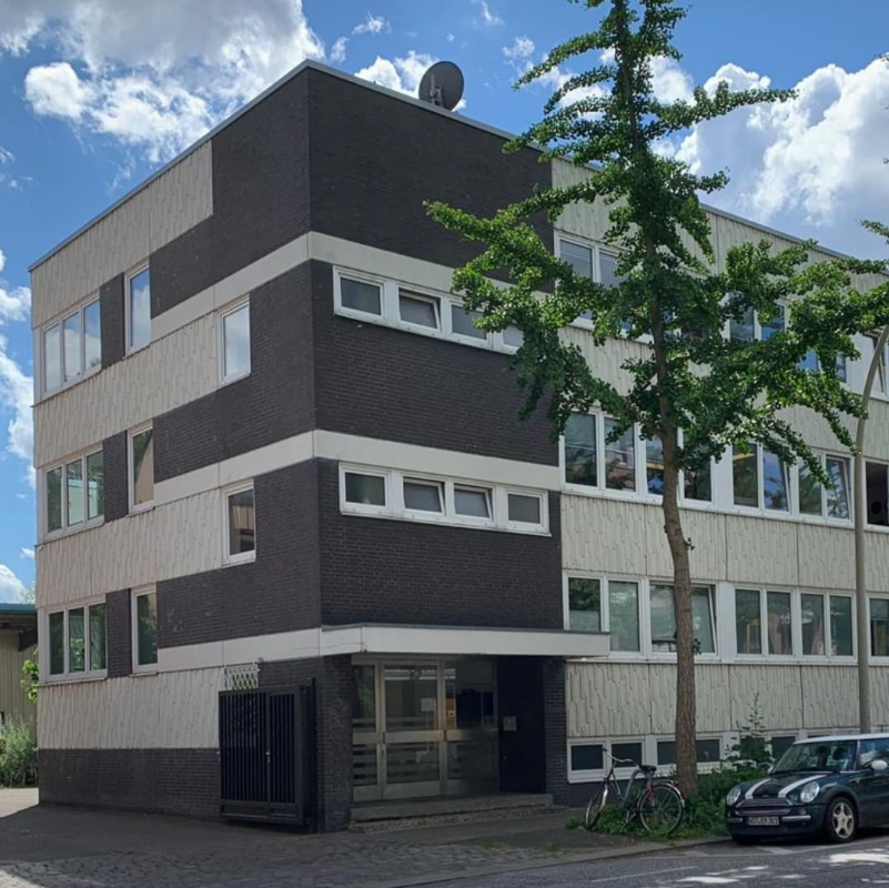 Dreistöckiges, grau-weißes Gebäude mit kleinen Fenstern. Davor stehen ein Baum und ein Auto.