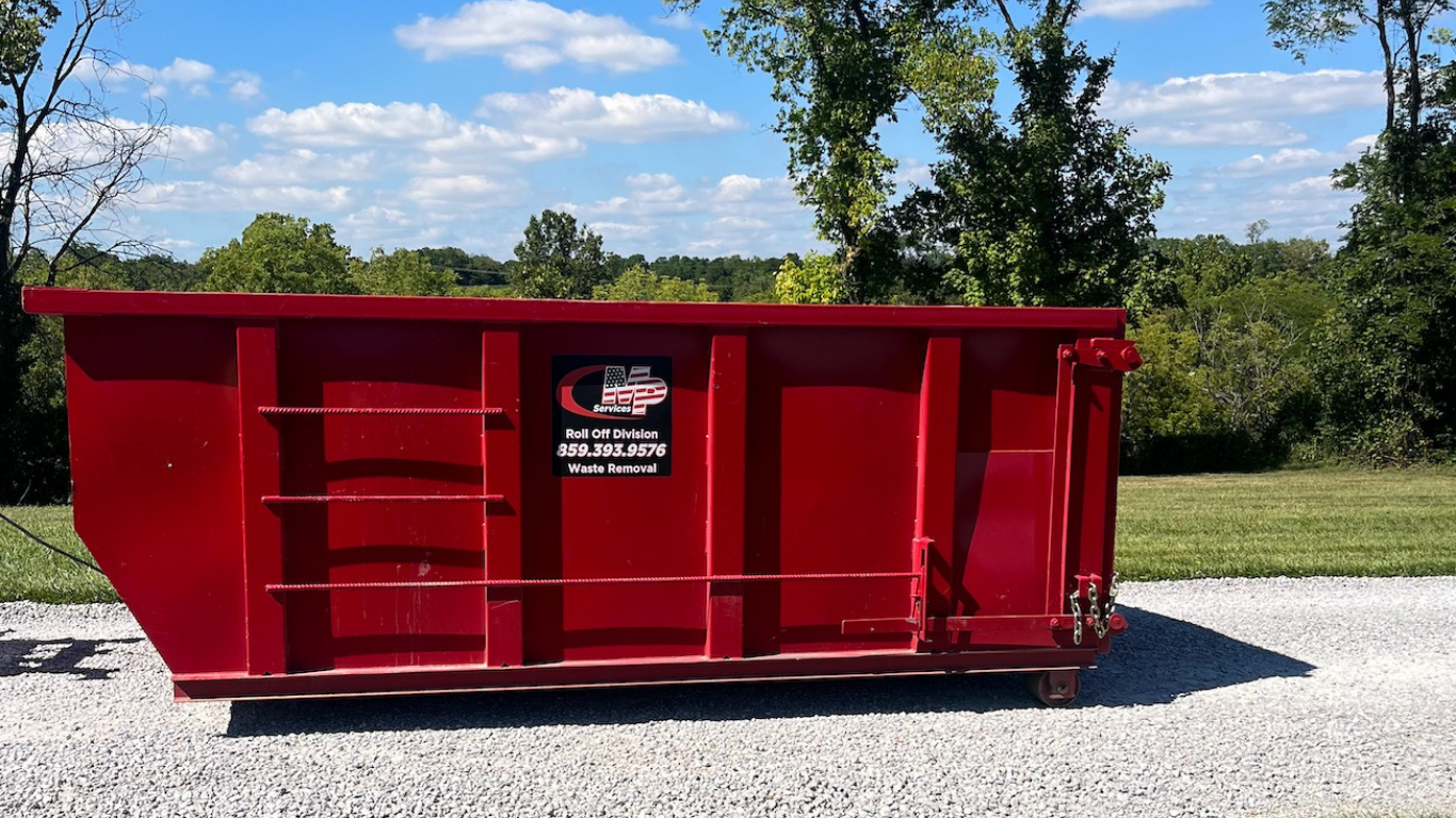 Red dumpster on gravel road, outdoors, trees in background.