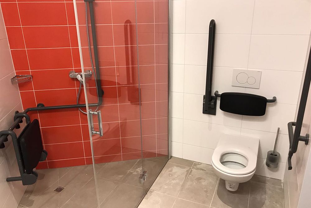 Links befindet sich eine barrierefreie Toilettenkabine mit WC, Haltegriffen und einem rot gefliesten Duschbereich.