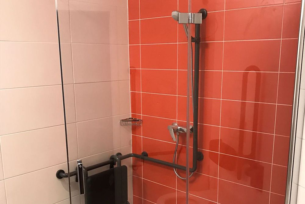 Barrierefreie Dusche mit Haltegriffen, Handbrause und schwarzem Handtuchhalter vor rot-weißen Fliesen