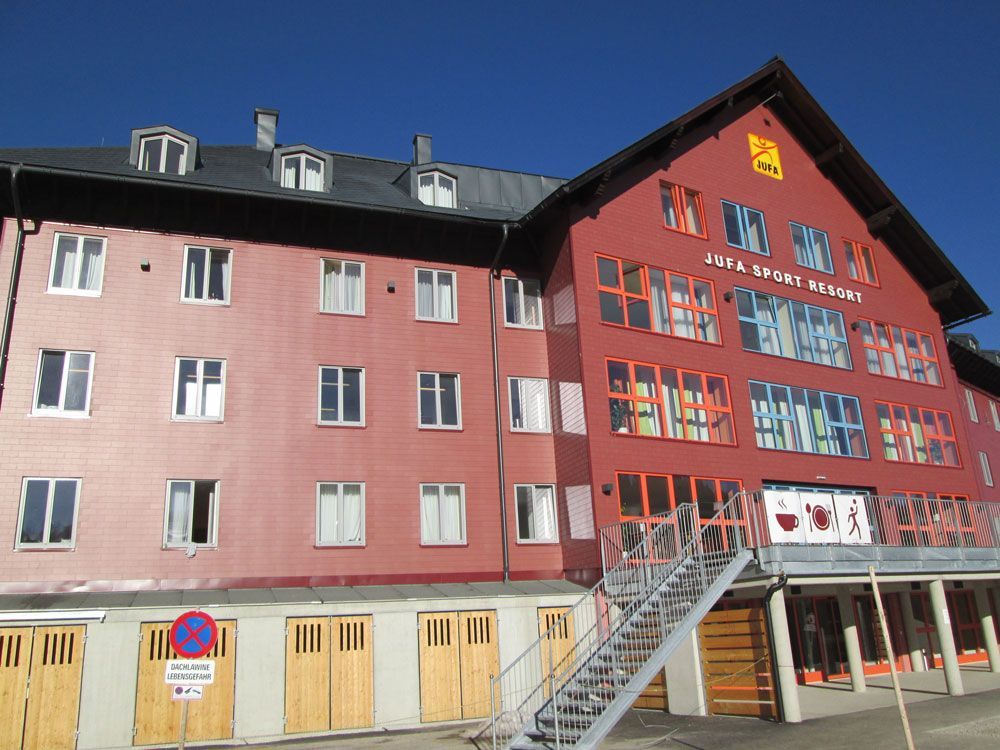 Rotes mehrstöckiges Gebäude mit dunklem Dach, vielen Fenstern und einer Außentreppe auf der rechten Seite.