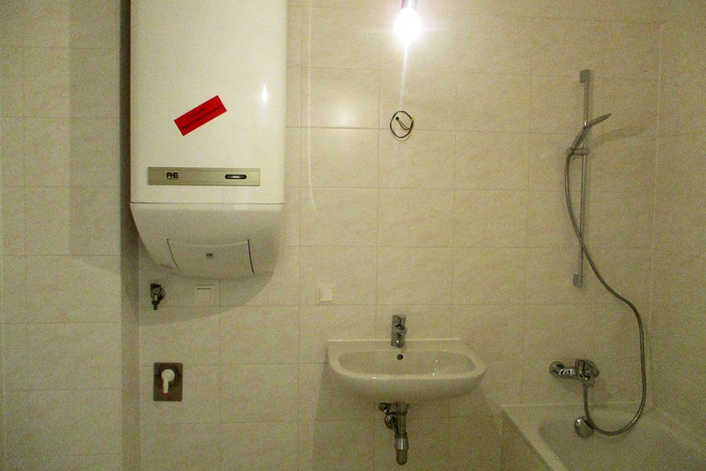 Barrierefreie Toilette mit wandmontiertem Waschbecken, Duschkopf und weißem Warmwasserbereiter über dem Waschbecken.