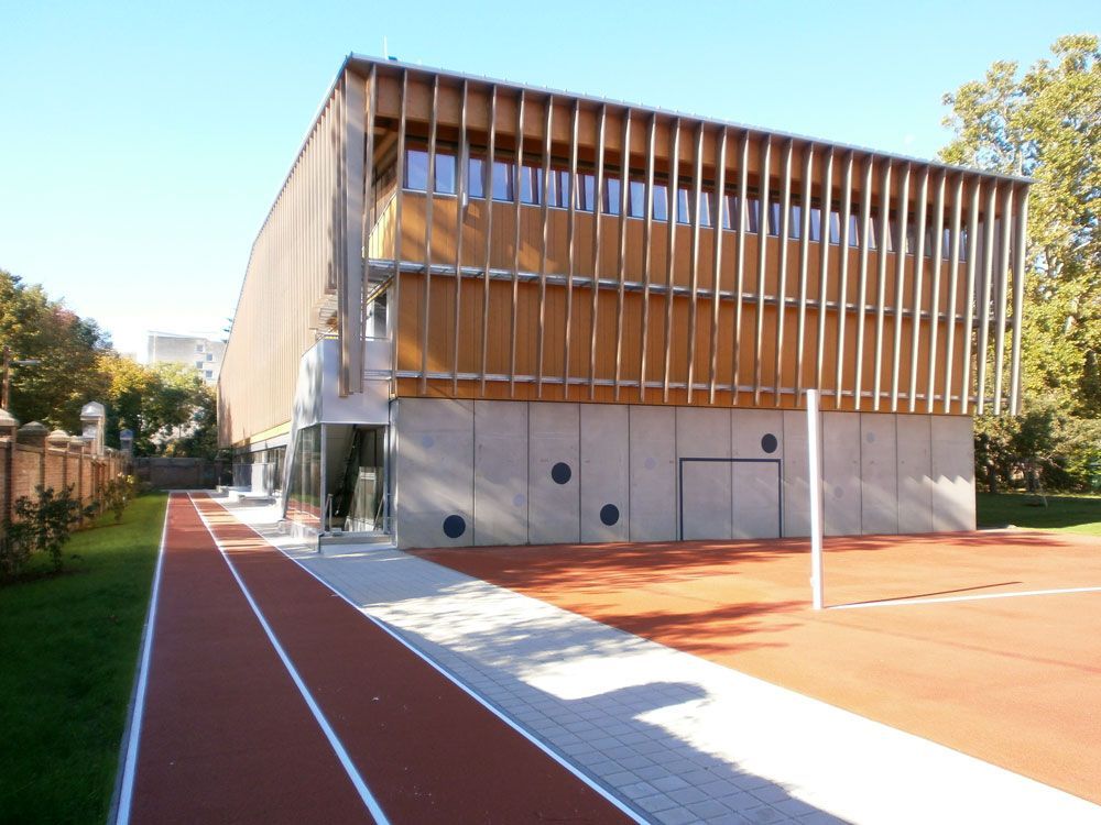 Modernes Gebäude mit Holzlatten, neben einer roten Laufbahn und einem offenen Hof unter klarem Himmel