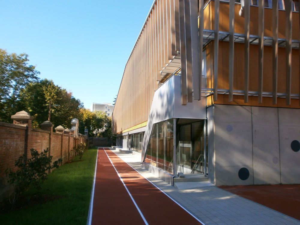 Modernes Gebäude mit gläsernem Eingang neben einer roten Bahntrasse und einer Backsteinmauer unter blauem Himmel