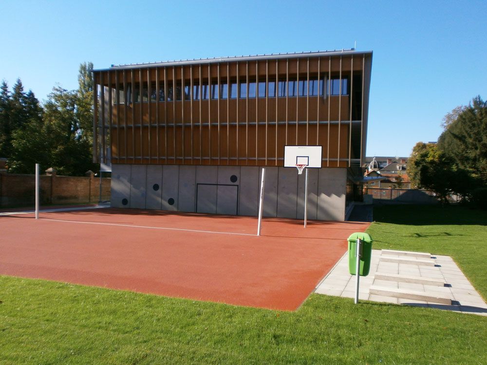 Freiluft-Sportplatz mit rotem Belag, Basketballkorb und einem modernen Holzgebäude im Hintergrund