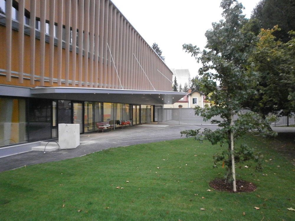 Modernes Gebäude mit überdachtem Gehweg, großen Fenstern und einer Rasenfläche mit einem kleinen Baum.